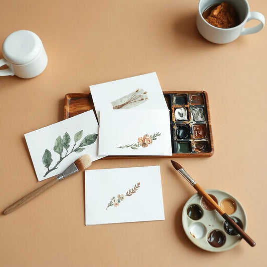 Hand-painted notecards with watercolor designs arranged beside brushes and a muted paint palette